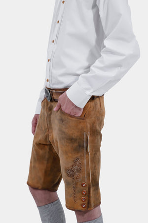 A man with a beard and styled hair wearing a white dress shirt and tan leather lederhosen shorts, standing with a slight side angle and his hand in his pocket, showcasing the lederhosen's detailed craftsmanship and grey knee-high socks.