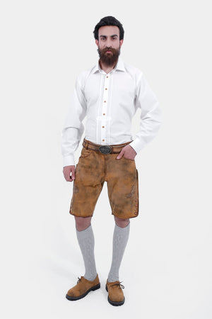 A man with a beard and styled hair wearing a white dress shirt paired with tan leather lederhosen shorts and grey knee-high socks, standing with his hands by his sides and looking directly at the camera.