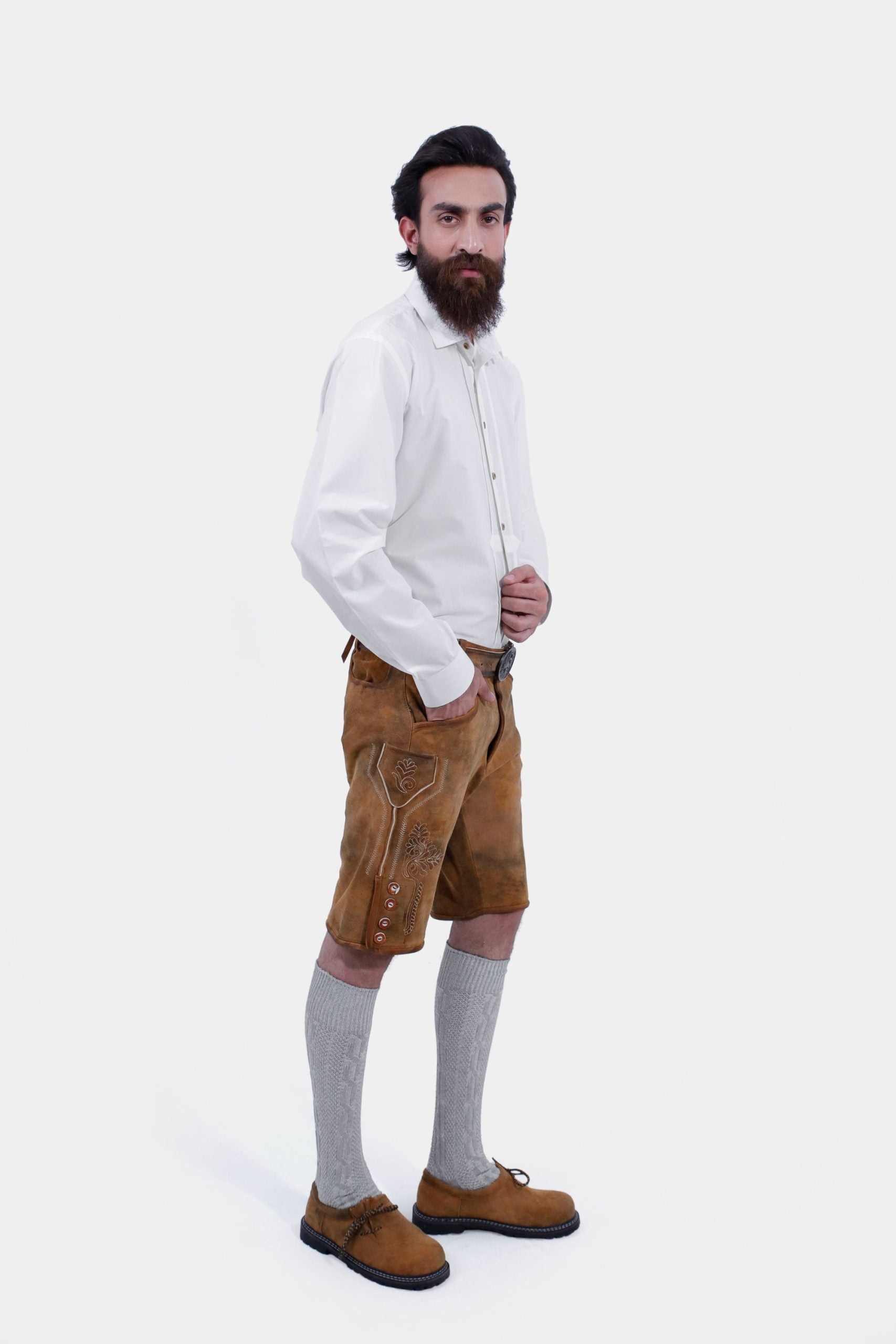 A man with a beard and styled hair wearing a white dress shirt and tan leather lederhosen shorts, standing with his hands in his pockets and looking directly at the camera, highlighting the intricate embroidery and button details.