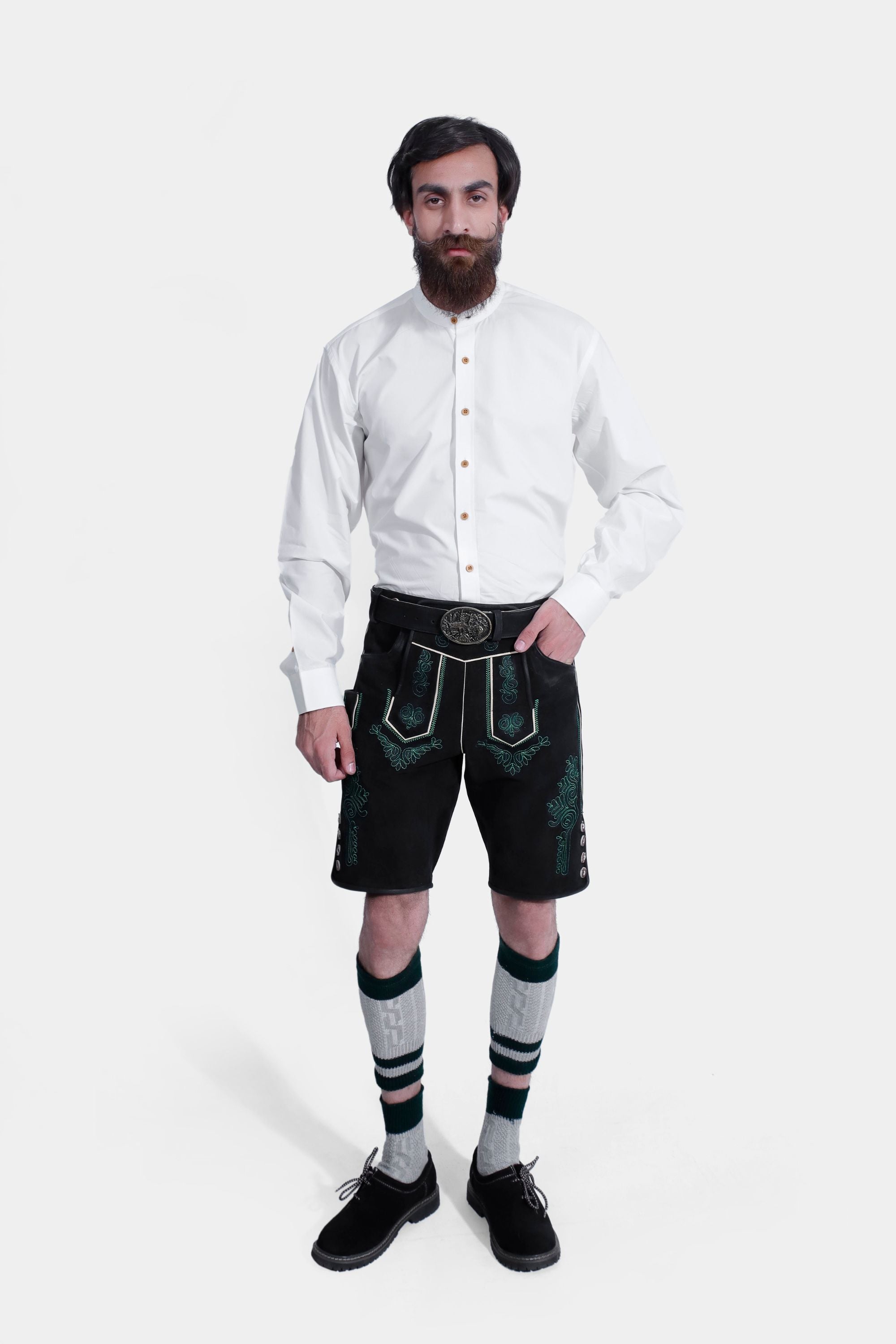 A man wearing a traditional Bavarian Lederhosen outfit, consisting of black knee-length Lederhosen with green embroidery, a white Bavarian shirt, knee-high socks, and black shoes, standing confidently with his hands in his pockets.