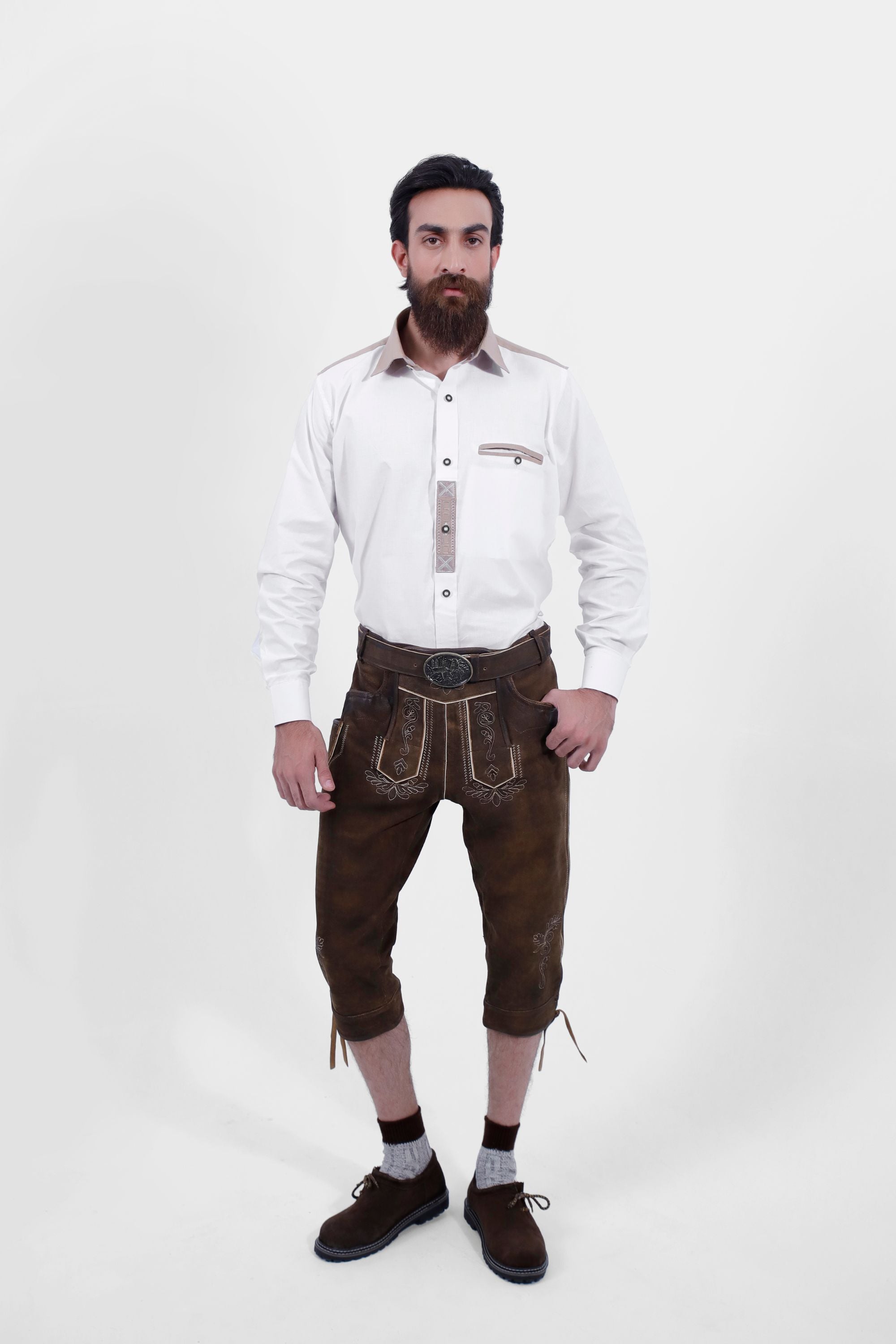 A bearded man stands against a white background wearing a white shirt and brown embroidered leather lederhosen with a decorative belt and dark brown shoes.