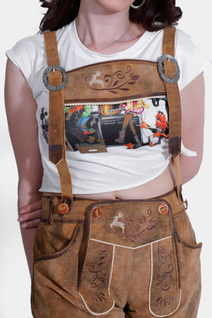 Royal Saxony Lederhosen Woman wearing tan lederhosen with suspenders over a white graphic T-shirt.