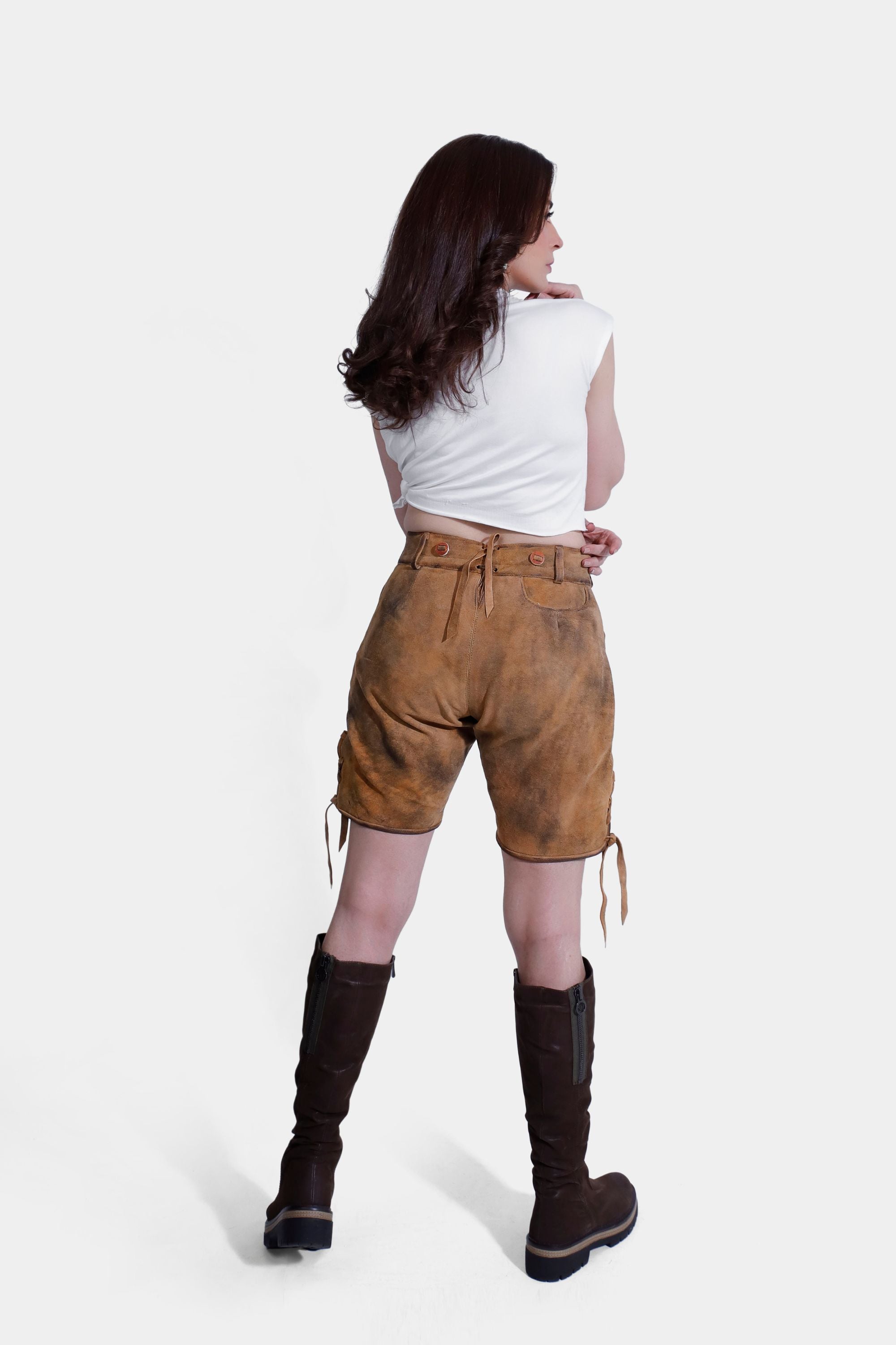 Back view of a Royal Saxony Lederhosen woman in tan lederhosen and a white T-shirt, with brown knee-high boots. Royal Saxony Lederhosen in Golden Amber Delight, goatskin leather with natural embroidery, brown horn buttons, and multiple pockets