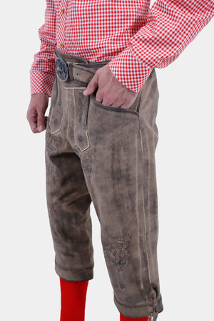 Side view of a man with a beard wearing a red and white checkered shirt, with his hand in the pocket of his light brown leather bundhosen, highlighting the intricate embroidery and pocket design, and bright red knee-high socks.