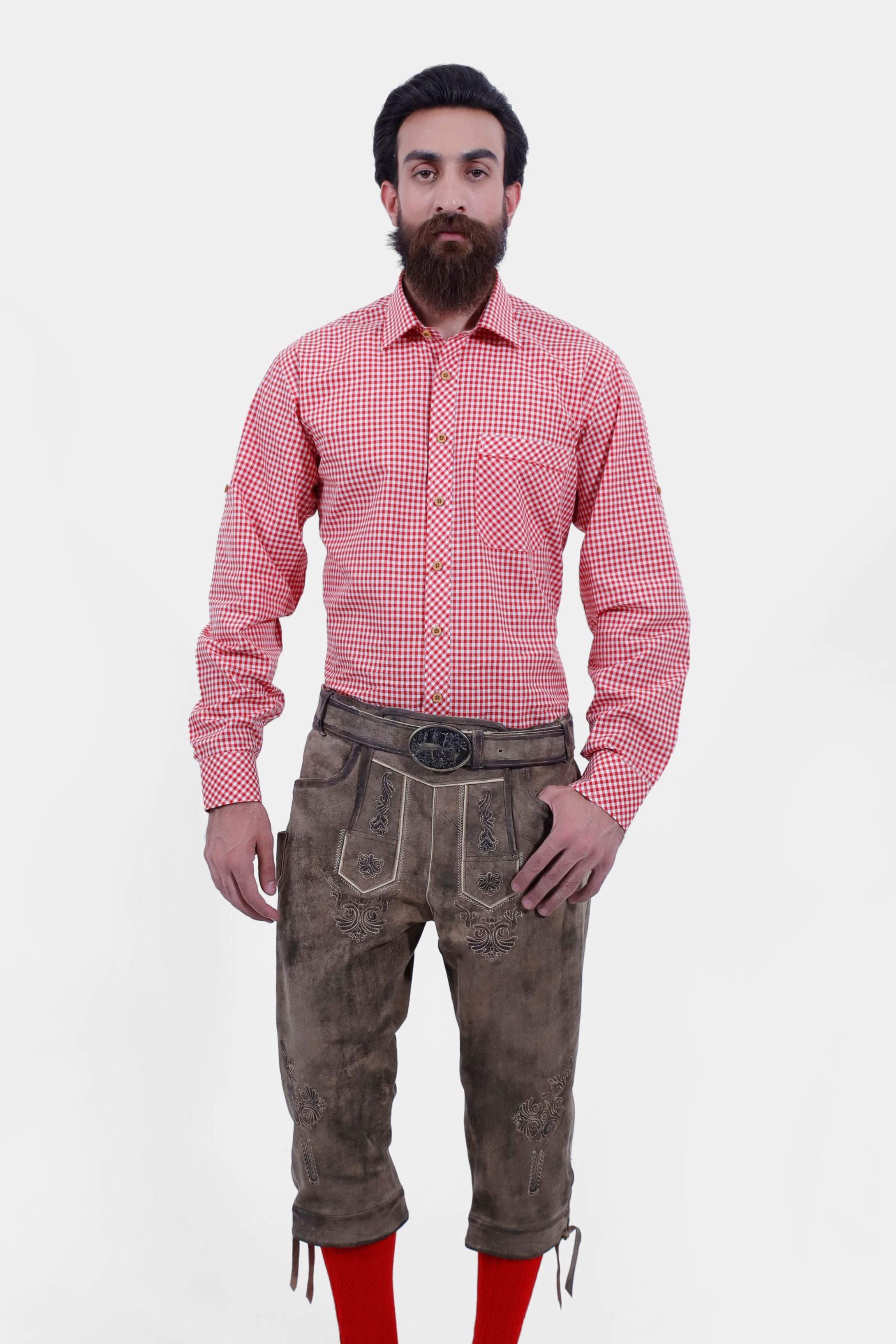 A man with a beard and styled hair wearing a red and white checkered shirt paired with light brown leather bundhosen and bright red knee-high socks, standing with his hands by his sides and looking directly at the camera.