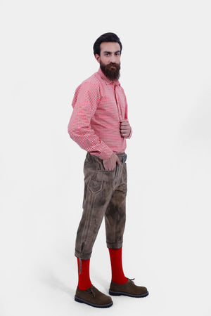 A man with a beard and styled hair wearing a red and white checkered shirt and light brown leather bundhosen, standing with a slight side angle and his hand in his pocket, showcasing the bundhosen's detailed craftsmanship and bright red knee-high socks.