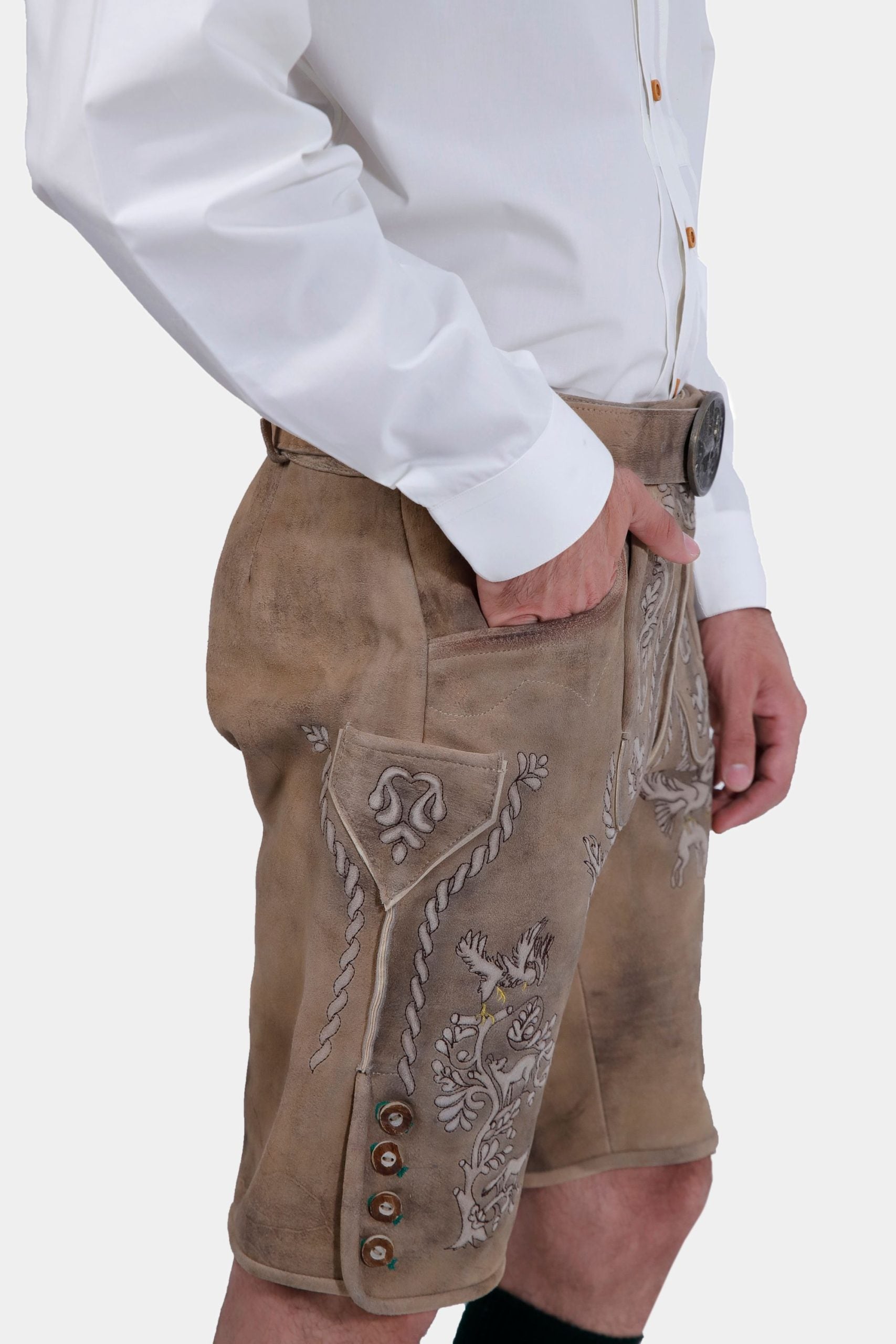 Side view of the man wearing the beige Lederhosen, showing the embroidery along the sides and the buttons on the lower part of the shorts. His hand is in his pocket.