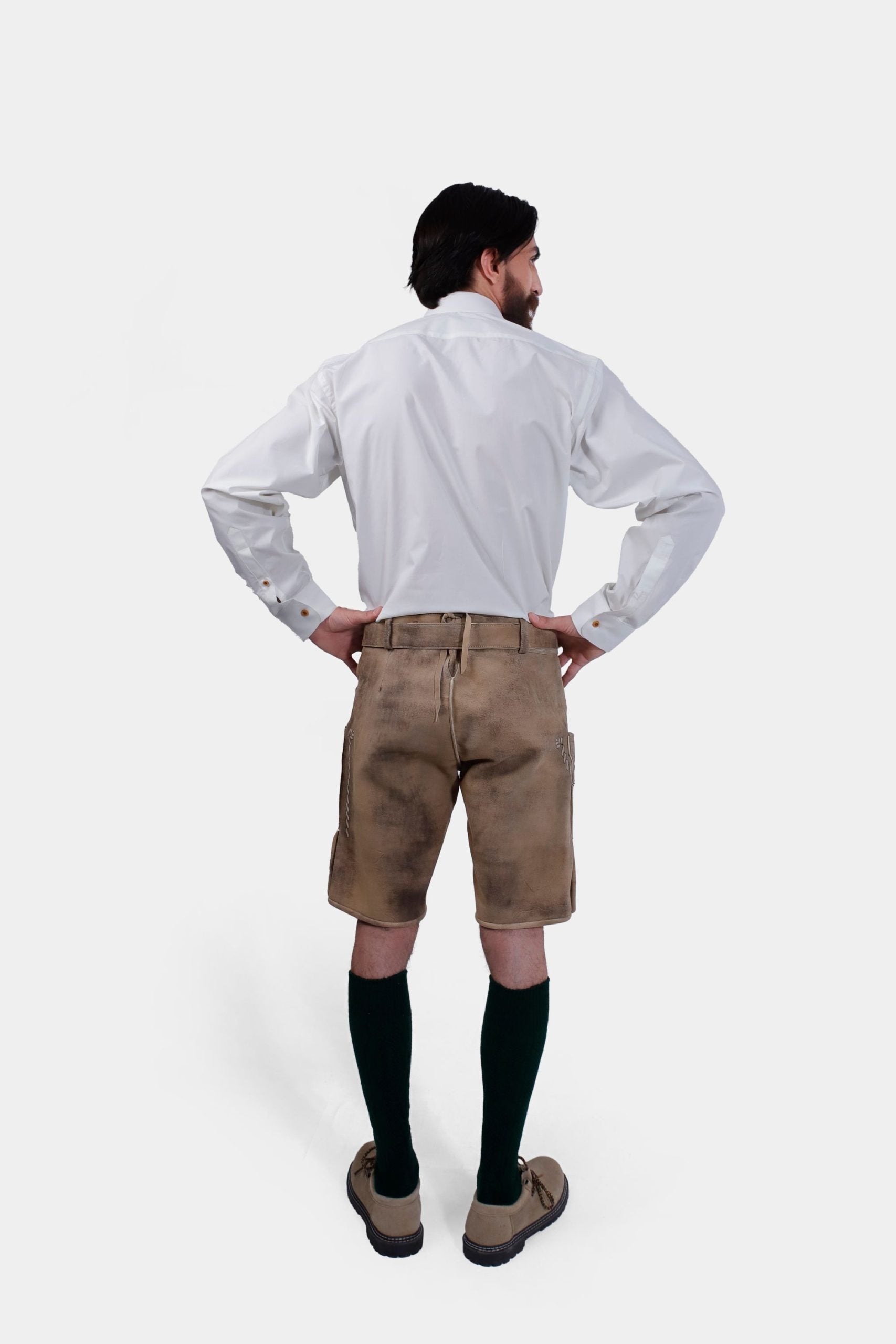 Rear view of the man showing the back of the beige Lederhosen and white shirt. His hands are on his hips, and the embroidery on the sides of the shorts is visible.