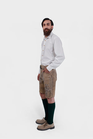 Side view of the man wearing the beige Lederhosen, showing the embroidery along the sides and the buttons on the lower part of the shorts. His hand is in his pocket.