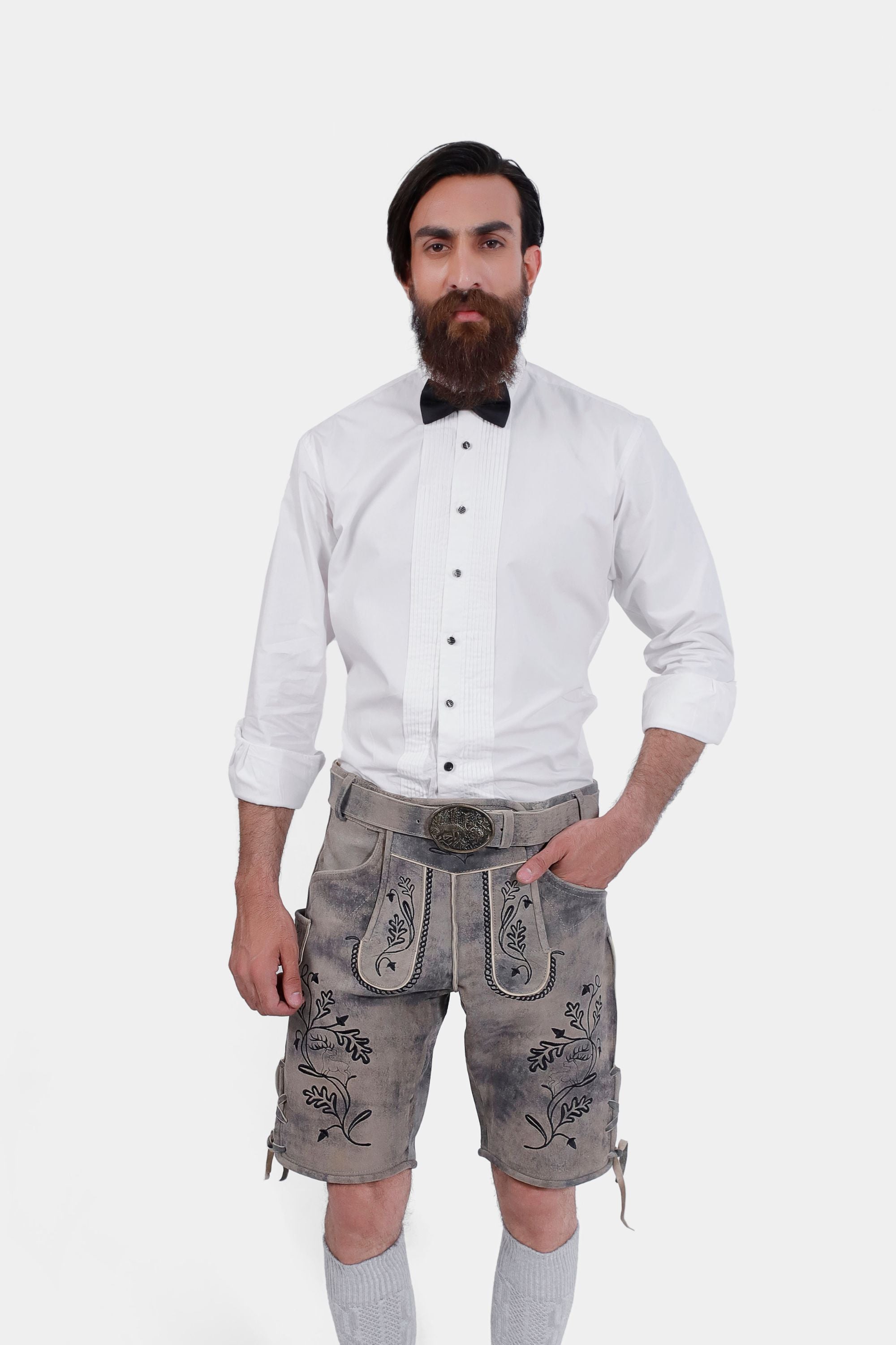 Man wearing Staffelsee Sunset Lederhosen in Oktoberfest Oak color, posing with hands in pockets and wearing a white shirt and bow tie