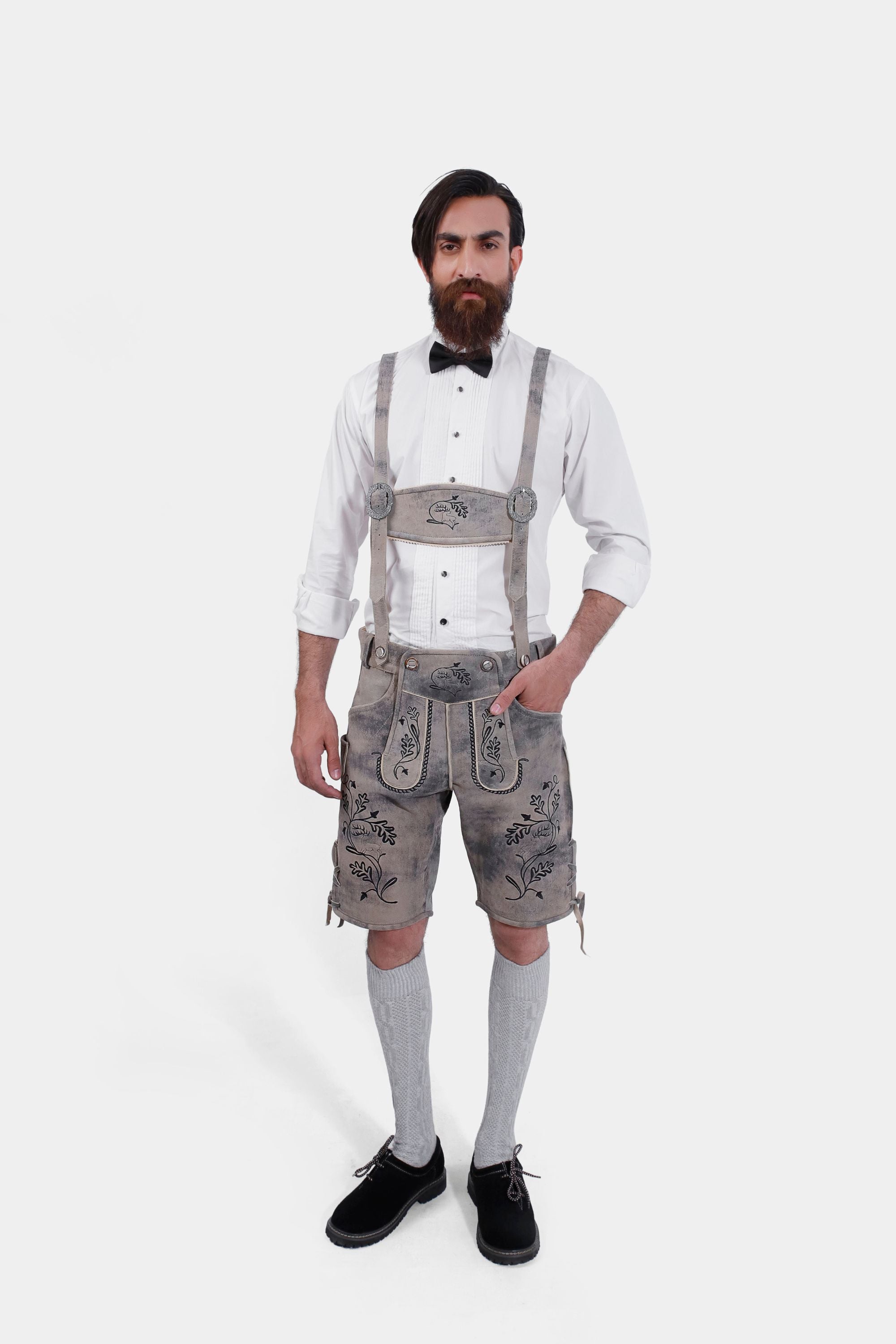 A man wearing traditional Bavarian Lederhosen. He is dressed in a white dress shirt with a black bow tie and light gray leather shorts adorned with intricate black embroidery and suspenders. He completes the outfit with light gray knee-high socks and black shoes, presenting a refined yet classic Bavarian style.