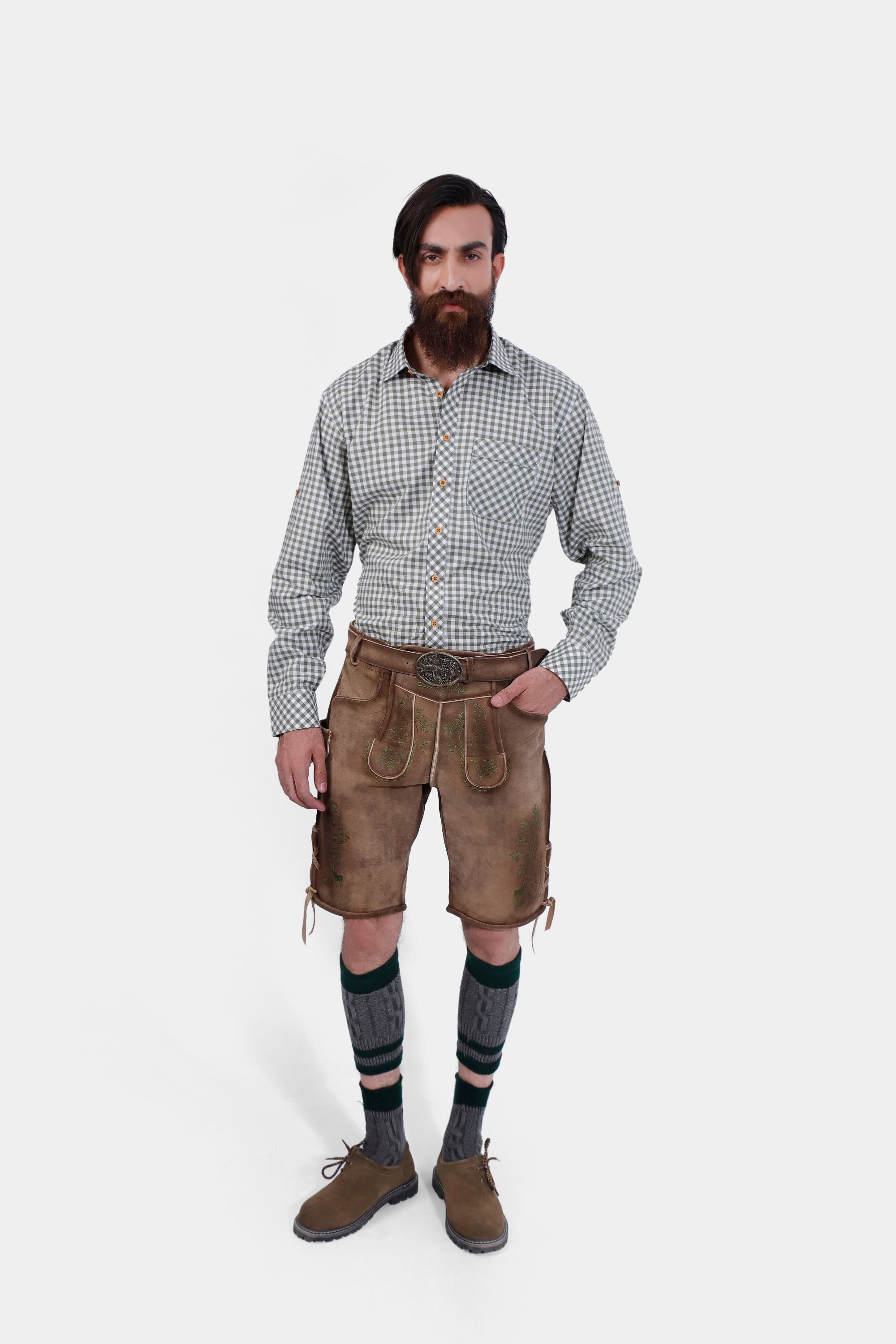 A front view of a man with a full beard wearing a gray and white checkered shirt and tan leather lederhosen with green embroidery, standing with both hands on his hips, showcasing the back design of the lederhosen.