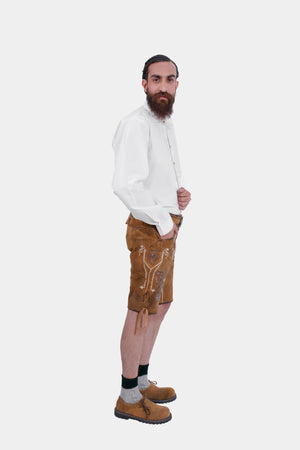 Side view of a man in a white dress shirt and brown lederhosen, showing detailed embroidery on the lederhosen. He has his left hand in his pocket and is looking slightly towards the camera.