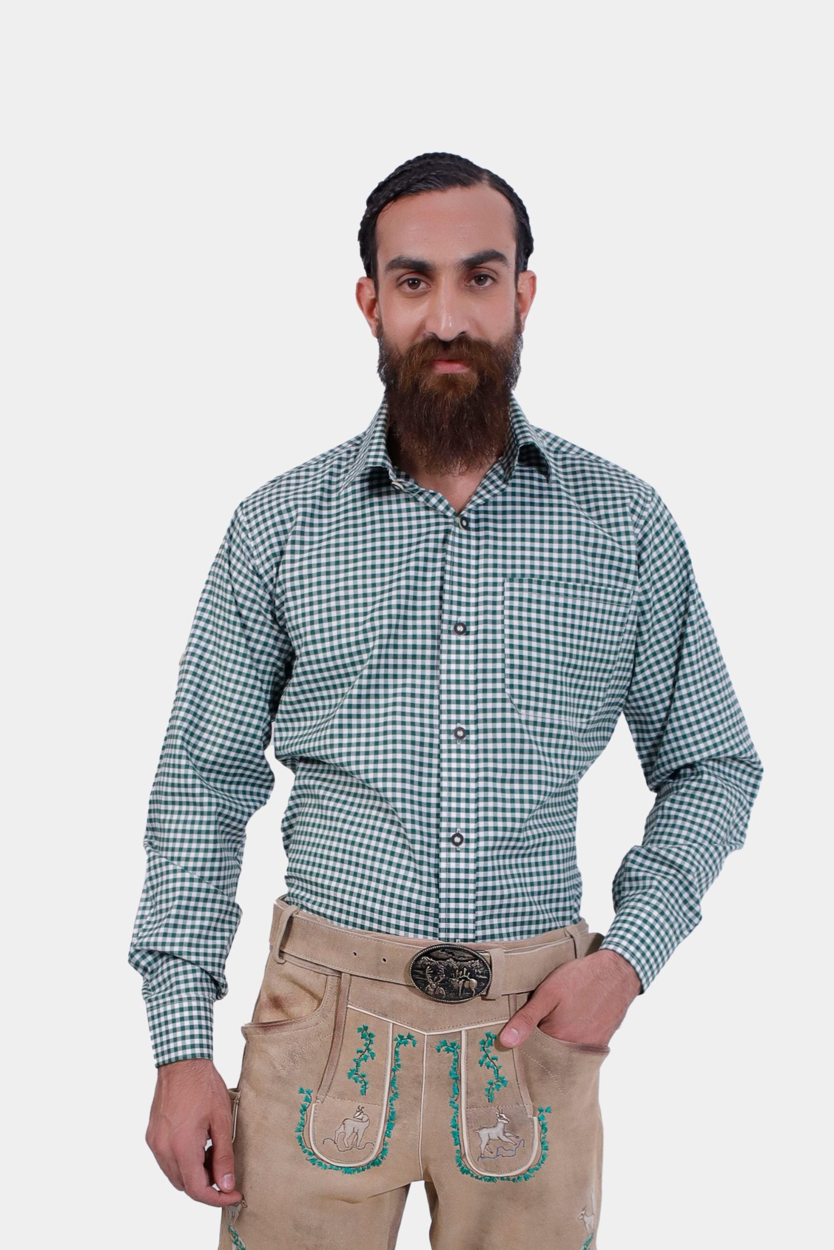 A bearded man wearing the Udo Kier Lederhosen Shirt, a green and white checkered shirt, paired with traditional beige lederhosen embroidered with green and yellow detailing. He stands confidently with one hand in his pocket against a plain white background. The shirt and lederhosen combination reflects classic Bavarian attire.