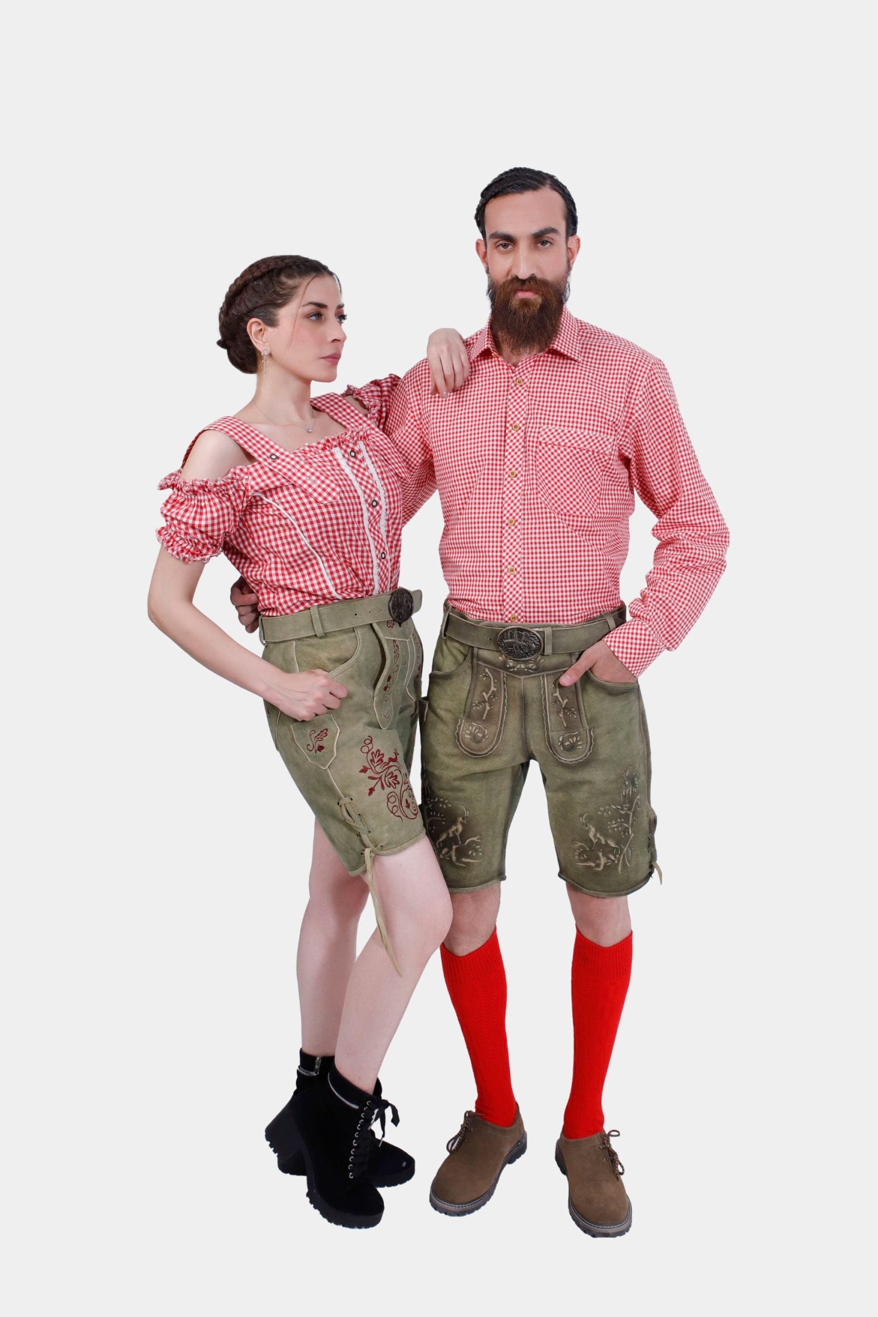 A couple posing in their coordinated Volksfest attire. The woman has her hand on the man's shoulder, showcasing her red checkered off-shoulder top and embroidered green leather shorts. The man stands confidently beside her in a matching red checkered shirt and embroidered green leather shorts, complete with red knee-high socks and brown shoes.