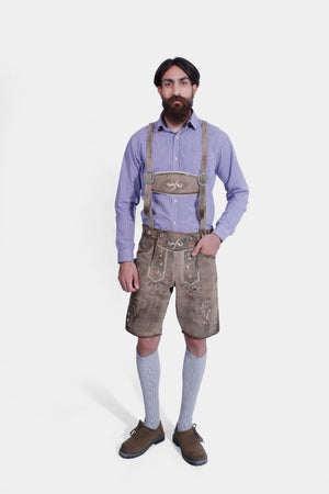 A man standing front-facing, wearing Stonewashed Fawn Lederhosen with suspenders, a blue checkered shirt, light grey knee-length socks, and brown shoes. His hands are in his pockets.