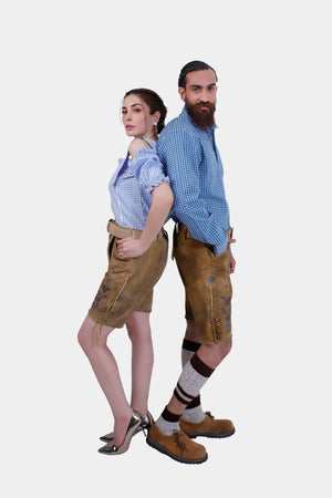 A couple wearing matching Wiesn outfits with intricately embroidered Lederhosen shorts, posing back to back. The man is dressed in a blue checkered shirt, and the woman is in a stylish off-shoulder blouse."