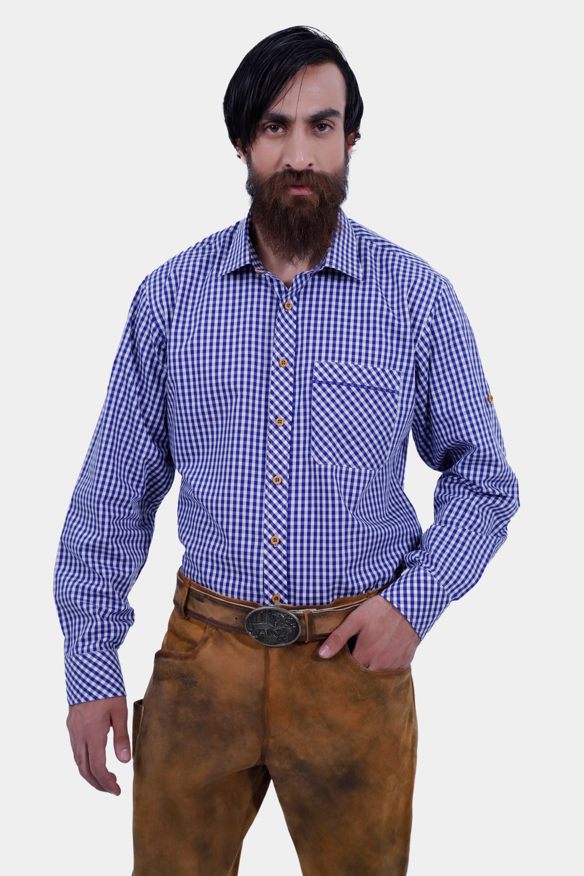 A man with a beard wearing a blue and white checkered shirt and brown leather long lederhosen. He stands with a neutral expression and hands in pockets, showcasing traditional alpine-style attire.