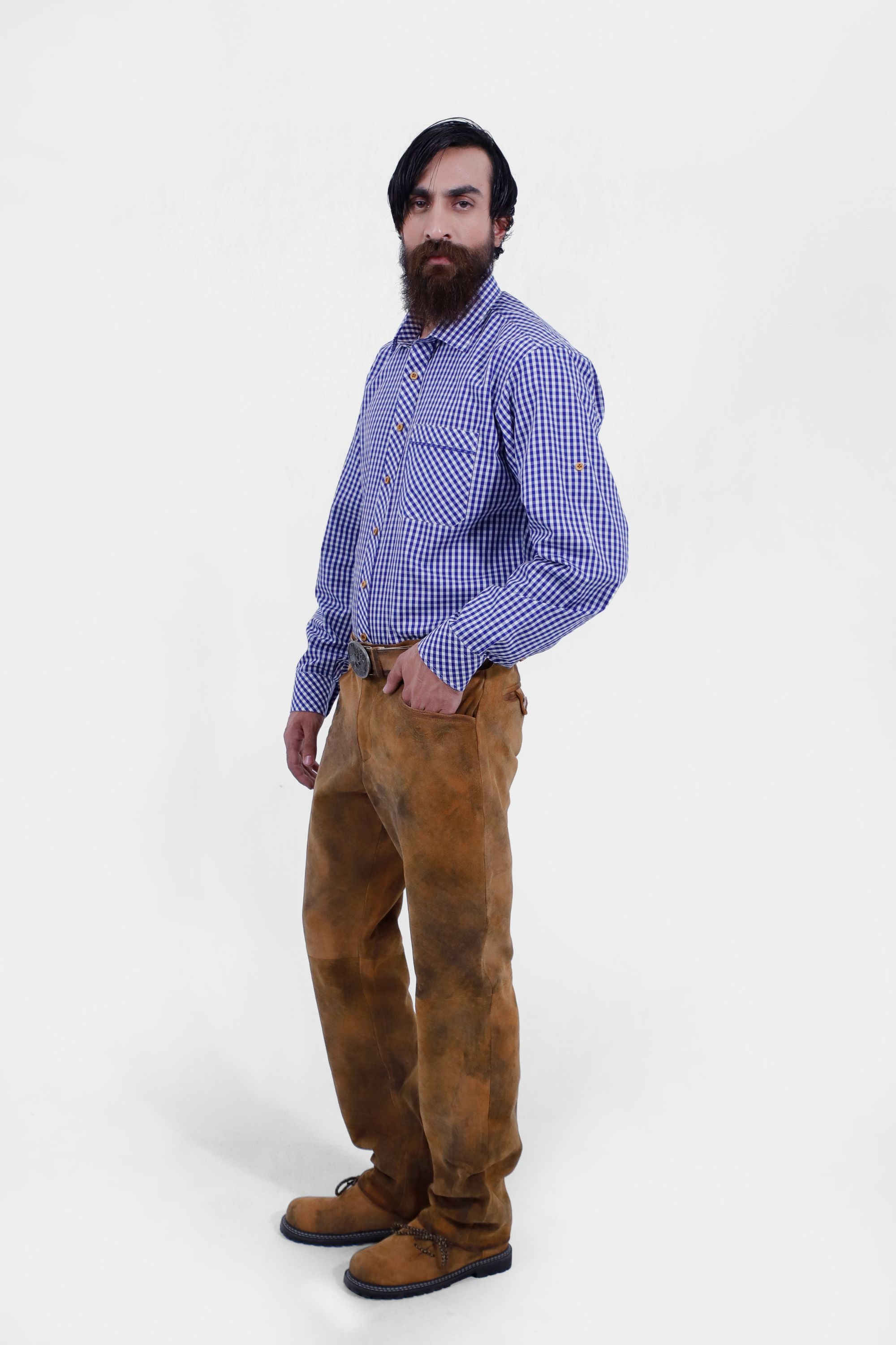 A man with a beard in a blue and white checkered shirt and brown leather long lederhosen, standing with one hand in his pocket. The image showcases the traditional alpine outfit from a side angle.