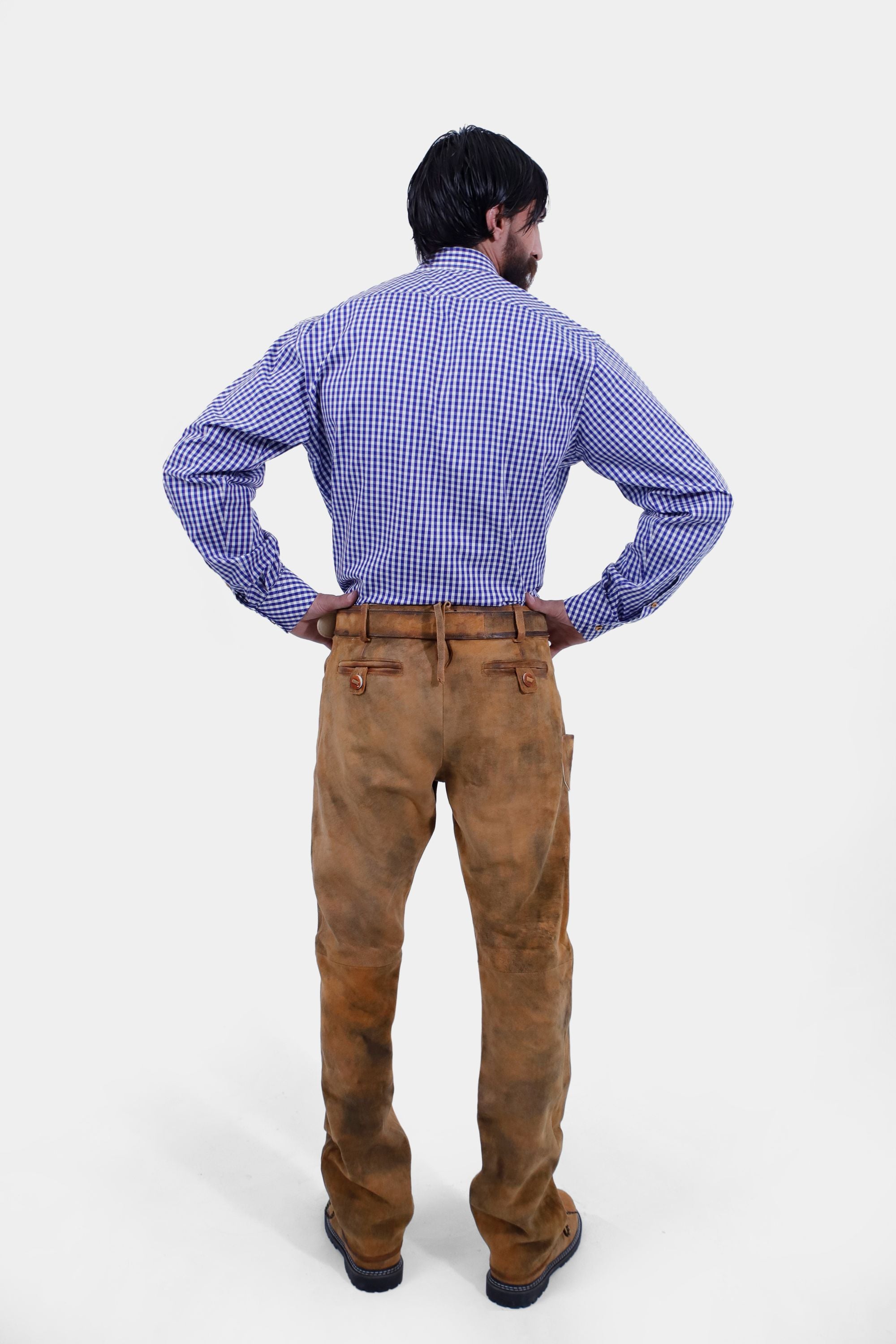 A full-length shot of a bearded man in a blue and white checkered shirt and brown leather long lederhosen, standing with his hands on his hips. The back view highlights the fit and details of the lederhosen.