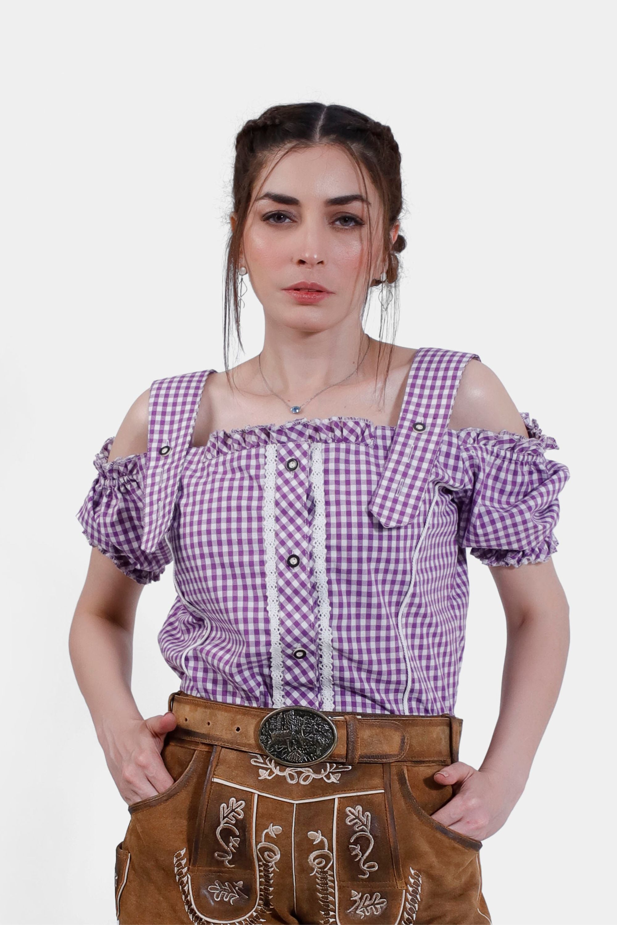 Woman wearing the Rihanna Traditional Blouse in purple checkered pattern, front view. The blouse features off-the-shoulder sleeves with ruffled edges and lace detailing along the buttons, paired with traditional leather trousers. The blouse showcases intricate design elements and provides a classic Trachten look.