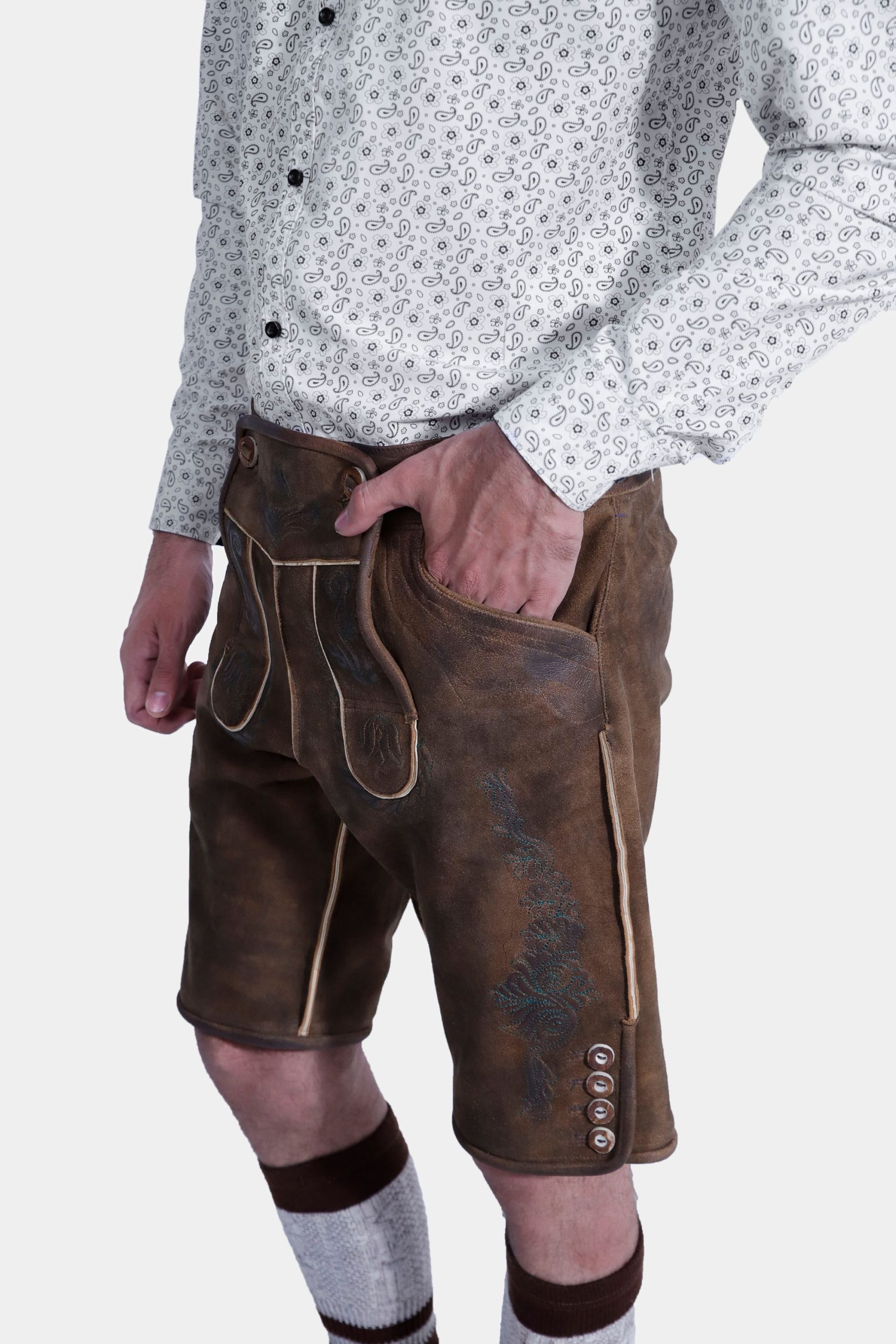 Closed side view of a man wearing traditional Bavarian Lederhosen. The outfit includes a white shirt with a subtle pattern and brown leather shorts with intricate embroidery. He completes the look with knee-high woolen socks and brown shoes. The attire captures the classic Bavarian style.