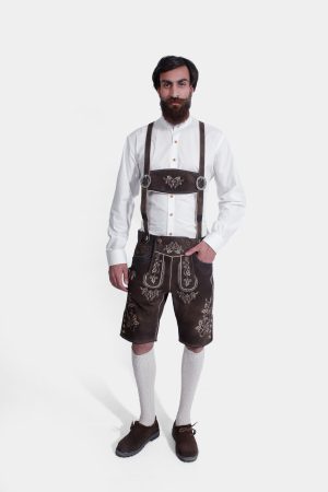 A man with a beard stands against a white background, wearing the "Angry Stag Eibsee Lederhosen." These traditional dark brown Lederhosen are knee-length and feature intricate light-colored embroidery on the bib, side panels, and around the leg openings. Complemented by a crisp white shirt, light grey knee-high socks, and dark brown shoes, his attire is a classic example of authentic Alpine fashion.