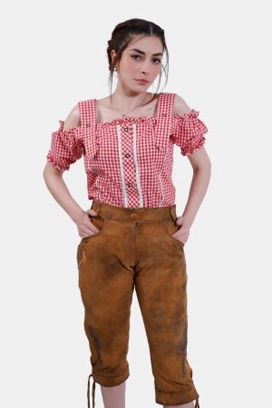 Front view of a woman wearing light brown knee-length Lederhosen with minimal embroidery and a red checkered blouse. Her hands rest in her pockets, and she looks directly at the camera.