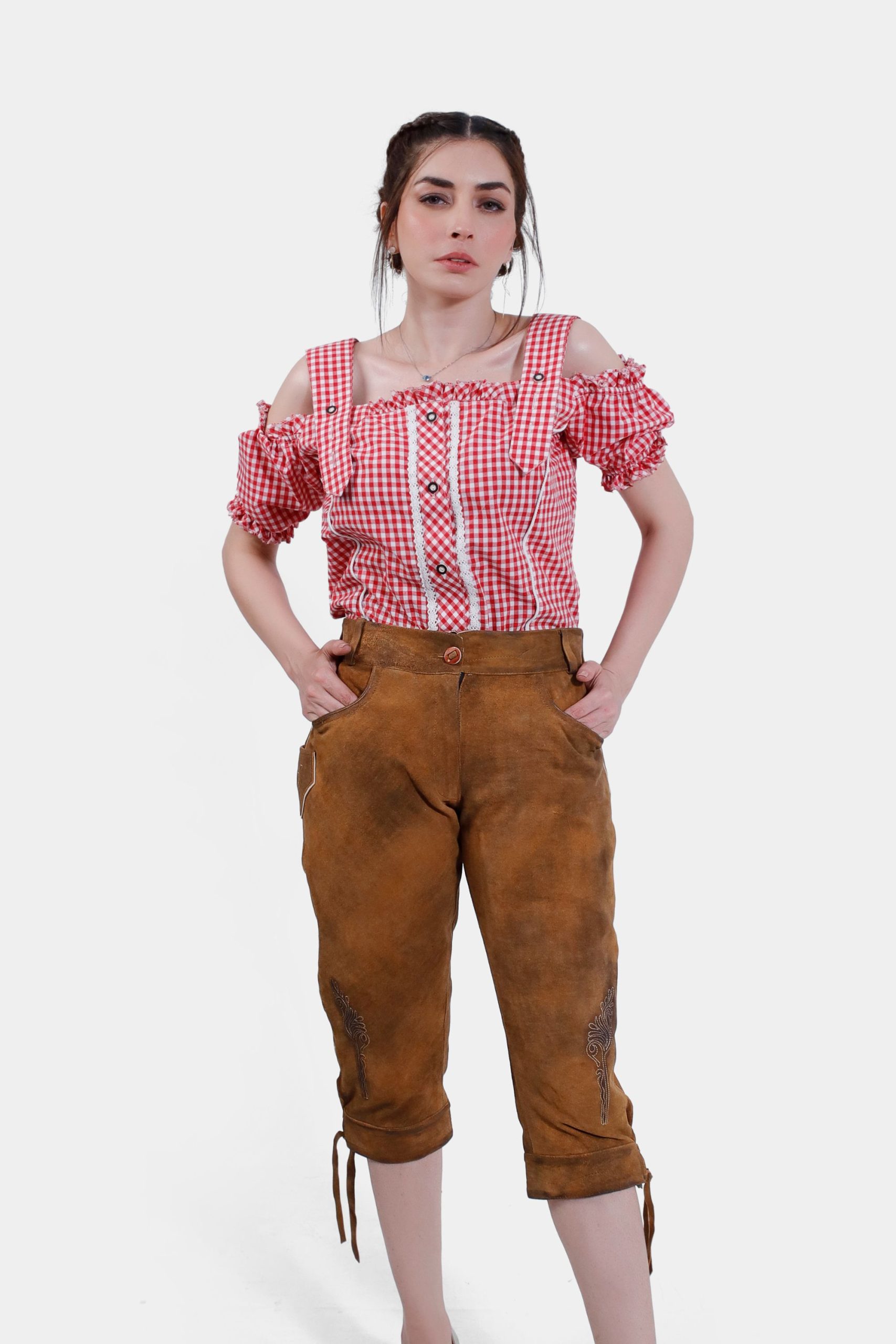A woman wearing a red checkered off-shoulder blouse paired with brown leather lederhosen. She stands confidently with her hands in her pockets, showcasing the detailed embroidery on the lederhosen.