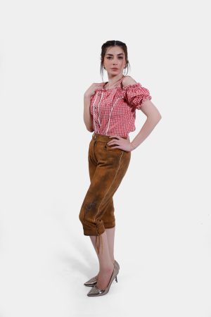 Full-body image of a woman wearing light brown knee-length Lederhosen with minimal embroidery, paired with a red checkered blouse. She stands confidently with one hand resting on her hip.