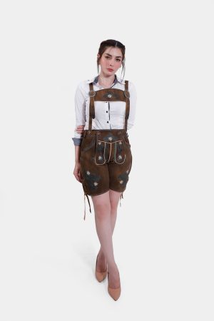A full-body shot of a woman wearing earthy brown lederhosen with green and white floral embroidery, paired with a white shirt and beige heels, standing with her hands on her hips.