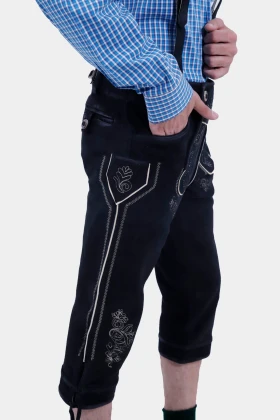 A side view of the man wearing a blue checkered shirt and black Bundhosen. The Bundhosen feature elaborate embroidery on the sides and front, with the man's hand resting in his pocket.
