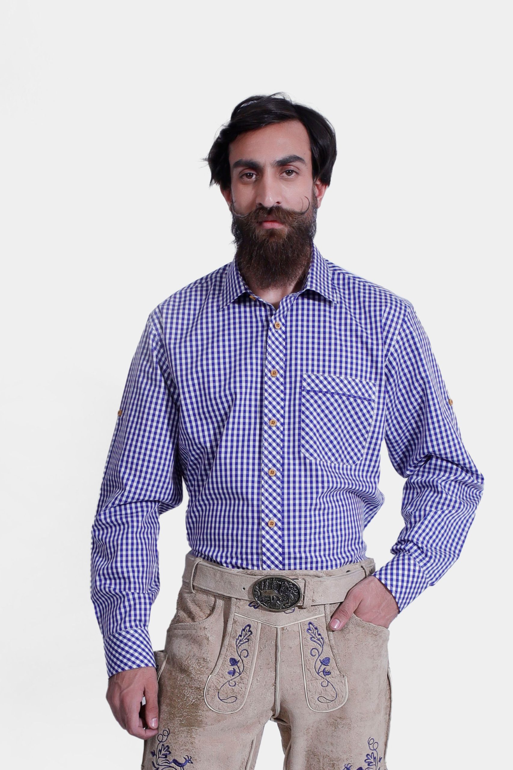 A man wearing the Bach Lederhosen Shirt, a blue and white checkered long-sleeve shirt with a front pocket. He pairs it with light brown leather Lederhosen adorned with blue embroidery and a decorative belt buckle. This traditional Bavarian outfit is perfect for Oktoberfest and cultural events, offering a vibrant and authentic look.