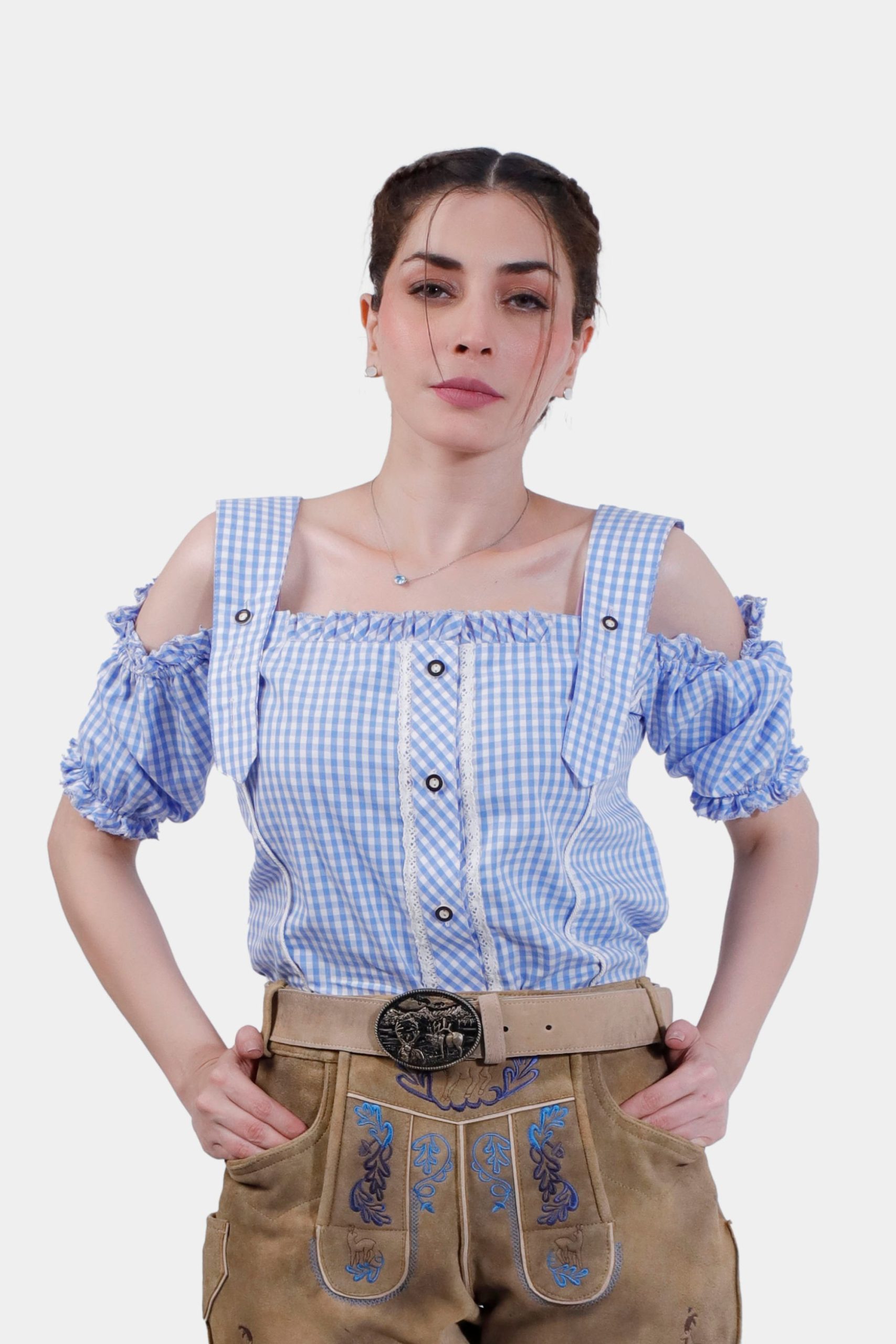 A woman wearing a blue checkered off-shoulder traditional blouse with ruffled sleeves and button details. She pairs the blouse with tan leather lederhosen featuring intricate blue embroidery and a decorative belt buckle.