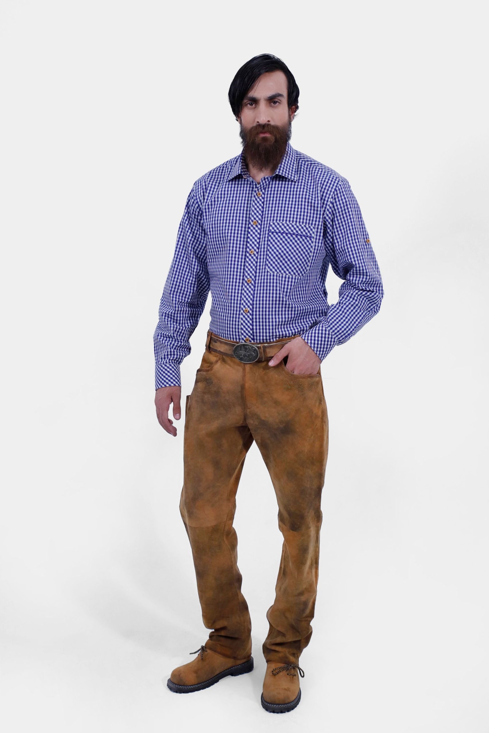 A man with a beard and long hair wearing a blue and white checkered shirt paired with brown leather lederhosen and brown boots, standing in a relaxed pose with his hands by his sides.