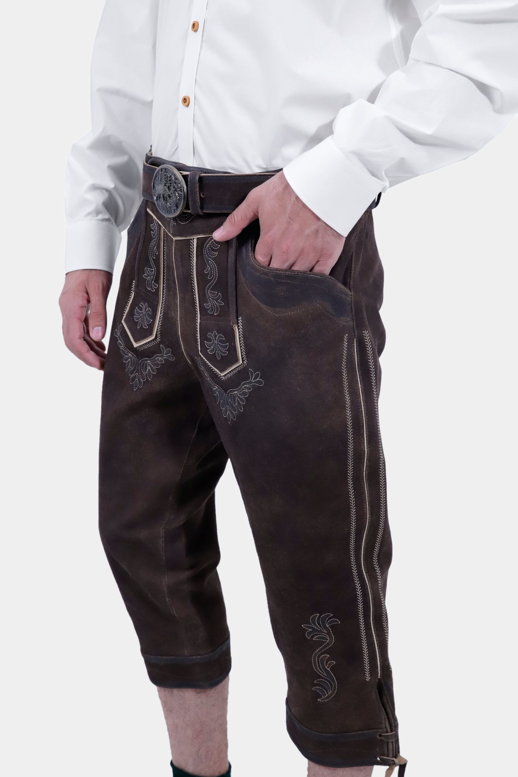 Side view of a man wearing traditional dark brown leather Bundhosen with intricate embroidery. The Bundhosen are paired with a white shirt and the outfit is accessorized with a leather belt and traditional shoes.