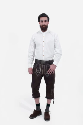 A man with a beard is wearing traditional Bavarian Bundhosen in a dark brown color. He is dressed in a white shirt with brown leather knee-length trousers adorned with intricate embroidery. The outfit is completed with traditional Bavarian shoes and socks.