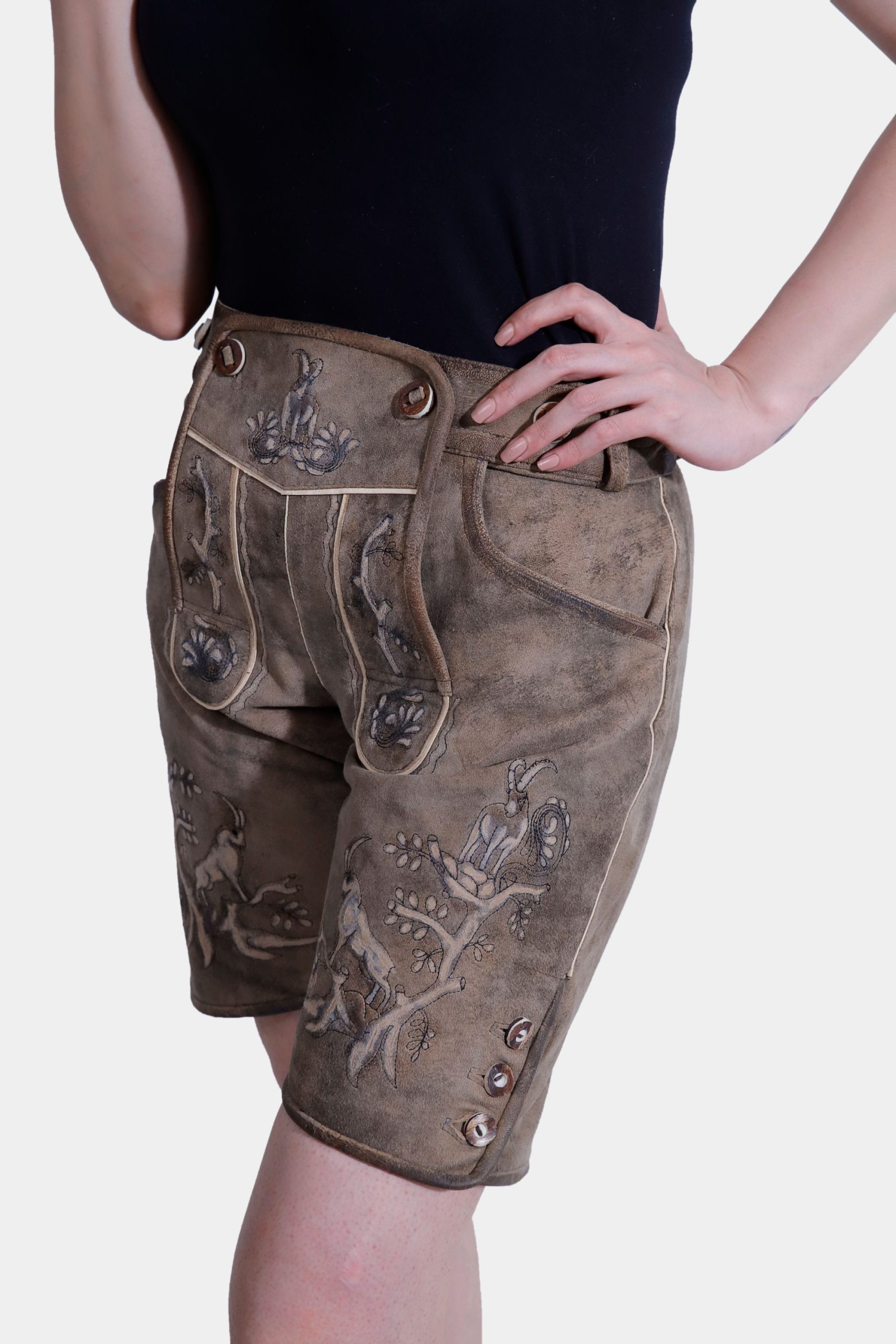 Side view of a woman in light brown Lederhosen with intricate embroidery and a pocket, paired with a black tank top. The image is set against a plain white background.