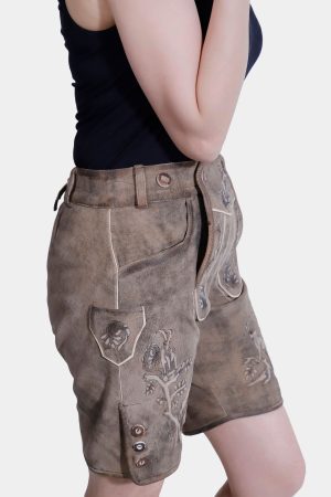 Side profile of a woman posing with one hand on her shoulder, wearing light brown Lederhosen with intricate embroidery. She is dressed in a black tank top, with a plain white background.