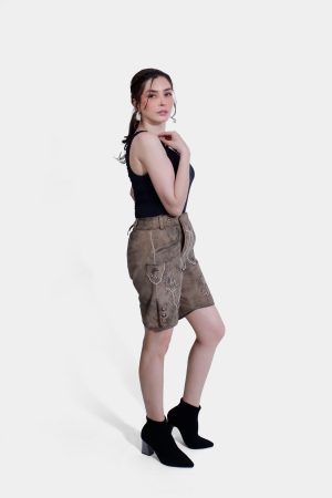 Side profile of a woman in light brown Lederhosen with intricate embroidery, paired with a black tank top and black ankle boots. The background is plain white.