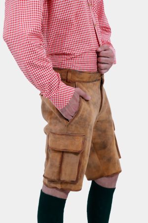 closed view of a man with a beard is standing in a three-quarter view against a white background, wearing a red and white checkered shirt and brown leather lederhosen. He has his right hand in the pocket of the lederhosen, and is also wearing dark green knee-high socks.