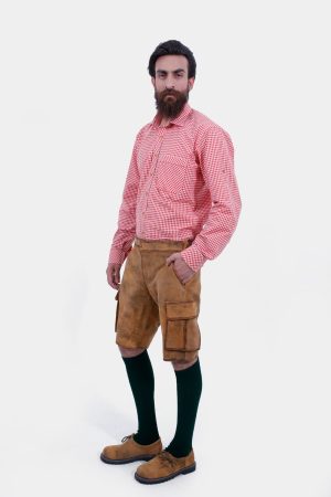 A man with a beard is standing in a three-quarter view against a white background, wearing a red and white checkered shirt and brown leather lederhosen. He has his right hand in the pocket of the lederhosen, and is also wearing dark green knee-high socks and brown shoes.