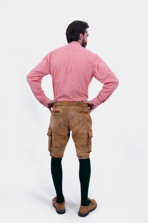 A man with a beard is standing with his back to the camera, hands on hips, wearing a red and white checkered shirt and brown leather lederhosen. He is also wearing dark green knee-high socks and brown shoes. The lederhosen has pockets on both sides and is detailed with stitching.