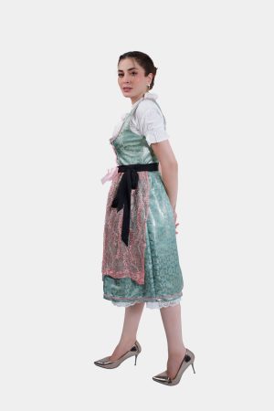 A side view of the woman in the dirndl, standing with her hand on her hip, looking slightly downward. The metallic heels add a modern touch to the traditional outfit.