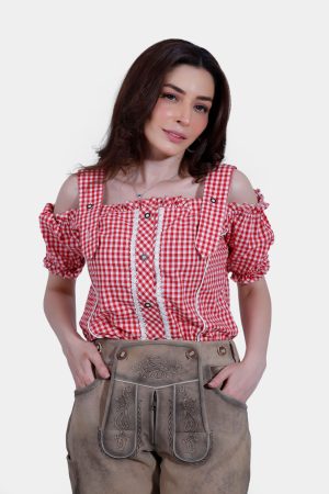 Close-up of a woman in a red and white checkered off-shoulder blouse and beige knee-length lederhosen. She has her hands in her pockets, highlighting the detailed embroidery and design of the lederhosen.