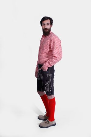 Side profile of a man wearing black Lahm Lederhosen with detailed white embroidery, paired with a red checkered shirt, red knee-high socks, and beige shoes, showcasing the traditional German attire
