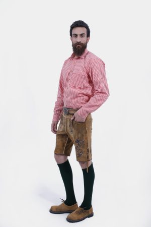 A man with a full beard wearing traditional Bavarian clothing, featuring a red and white checkered shirt and tan leather lederhosen with brown embroidery, paired with green knee-high socks and brown shoes, standing with one hand in his pocket.