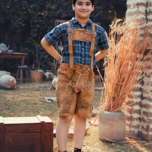 A young boy stands outdoors in a sunny garden, modeling the "Brajan Gruda Lederhosen Shorts." These traditional brown Lederhosen shorts feature natural colored embroidery on the bib and legs. He is wearing them with a plaid blue and green shirt, suspenders, and brown boots. The rustic scene includes a wooden box and tall dried grass beside a brick column, with lush greenery and flowering bushes in the background.