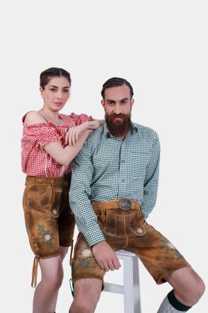 A couple wearing matching Freude Couple Lederhosen outfits. The man is seated wearing a green checkered shirt and Lederhosen shorts, while the woman stands beside him in a red checkered off-shoulder blouse and Lederhosen shorts.
