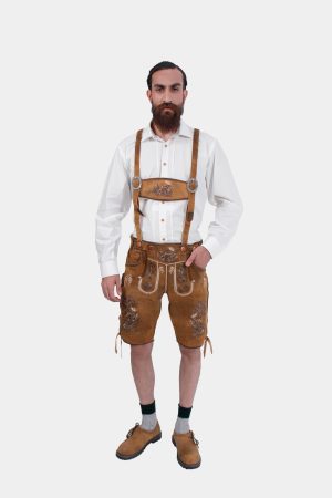 Full front view of a man wearing a white dress shirt and brown lederhosen with suspenders. The suspenders are decorated with intricate embroidery, and he has his left hand in his pocket.